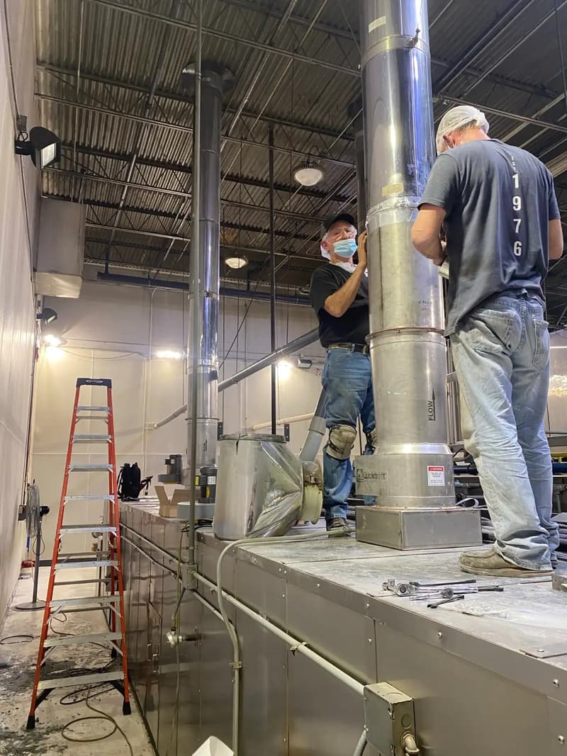 Two Henry Group technicians fitting a stainless exhaust stack to the top of a bakery oven