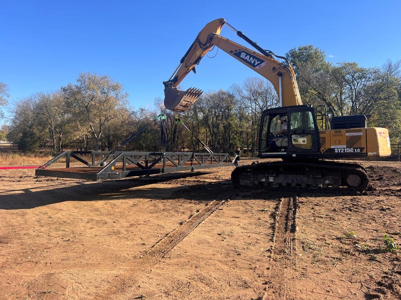 An excavator lifting a steel truss bridge into place on site