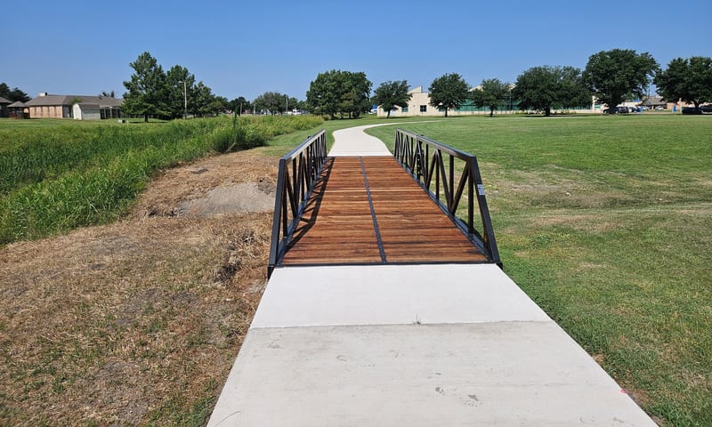 A Roadrunner steel trail bridge with a wood deck on a concrete park path